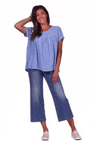 A woman wearing a blue crinkle linen gathered top with a round neckline, short sleeves, and displayed gathers at the bodice. She is paired with blue jeans and white shoes.