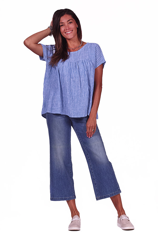 A woman wearing a blue crinkle linen gathered top with a round neckline, short sleeves, and displayed gathers at the bodice. She is paired with blue jeans and white shoes.